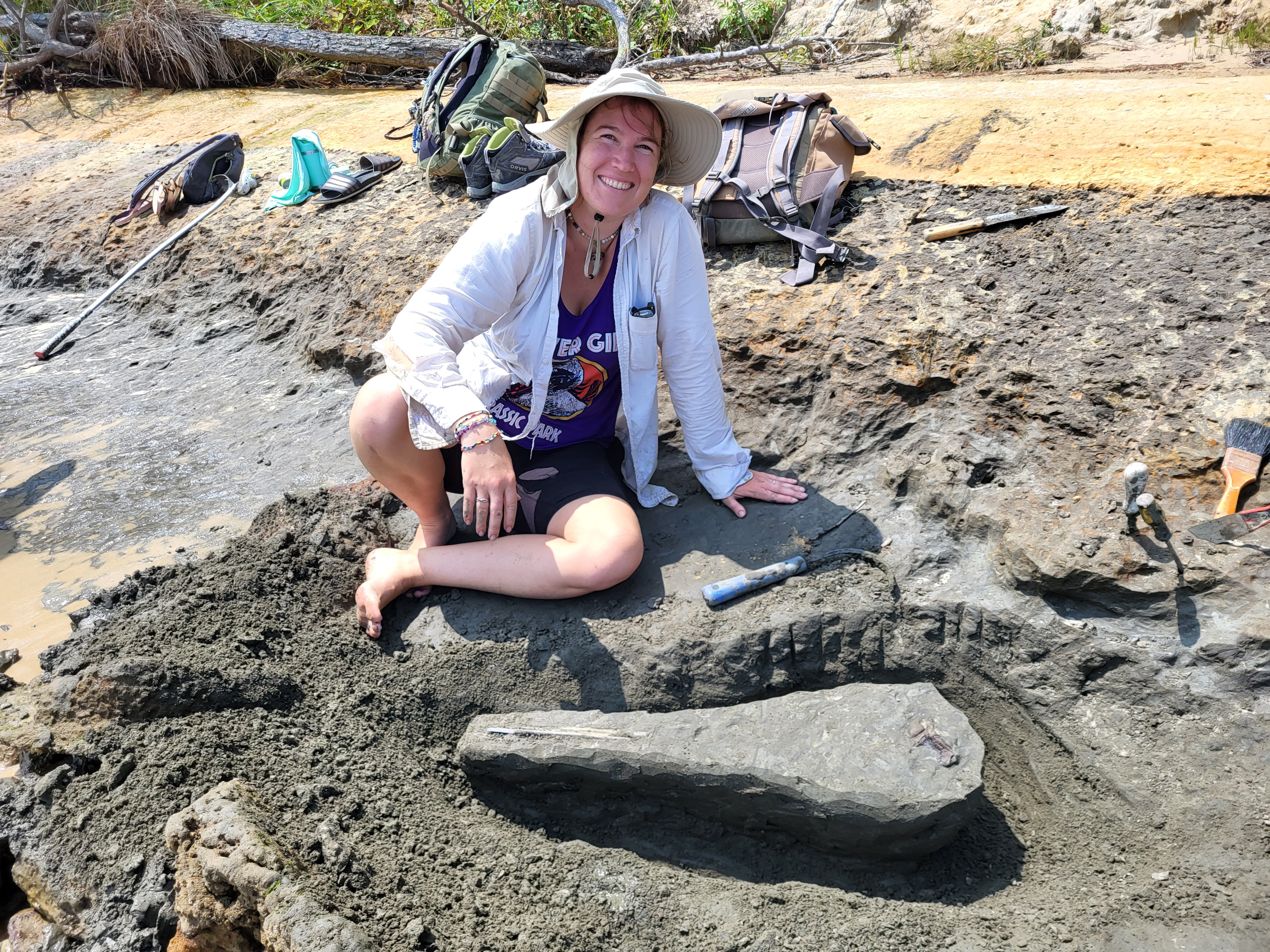 A smiling woman in a white shirt and hat sits on a beach with a partially excavated fossil nearby. Next to the fossil is a CobraHead Weeder.