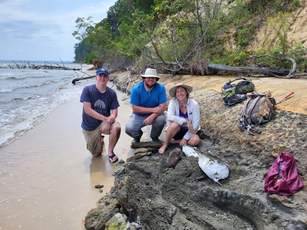 Three people pose in a crouching position on a beach near a partially excavated and encased fossil.